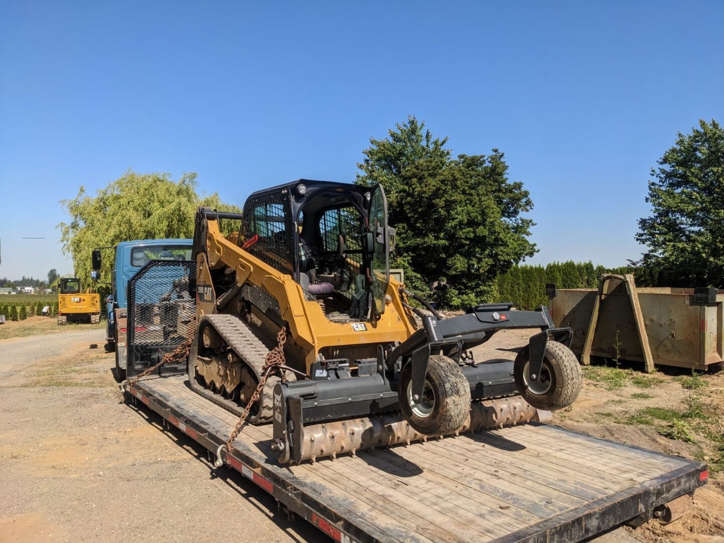 A bulldozer on a flatbed trailer parked on a dirt road, with clear skies and trees in the background.