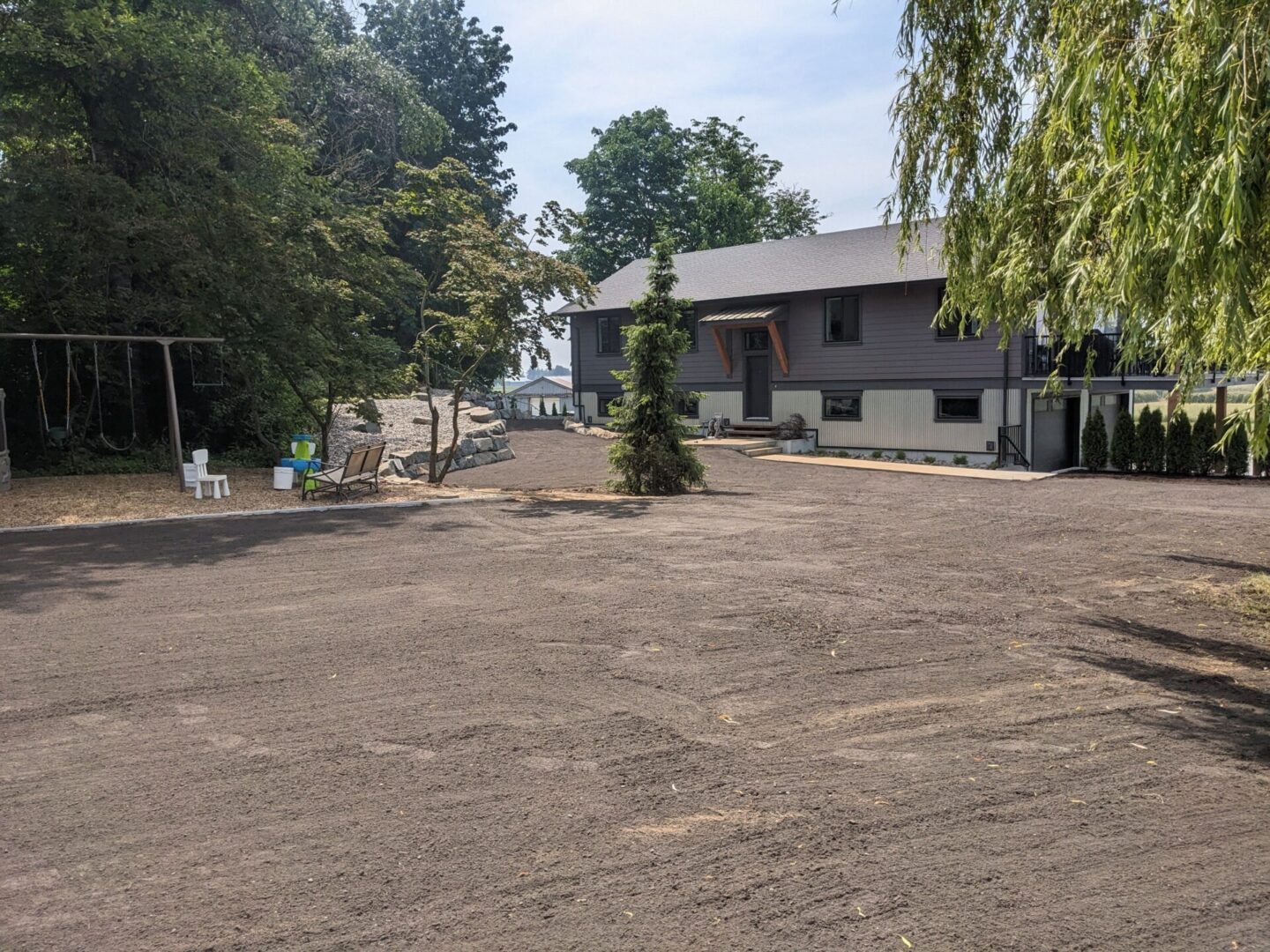 A newly paved driveway leading to a modern gray house with trees and a sandy area on a sunny day.