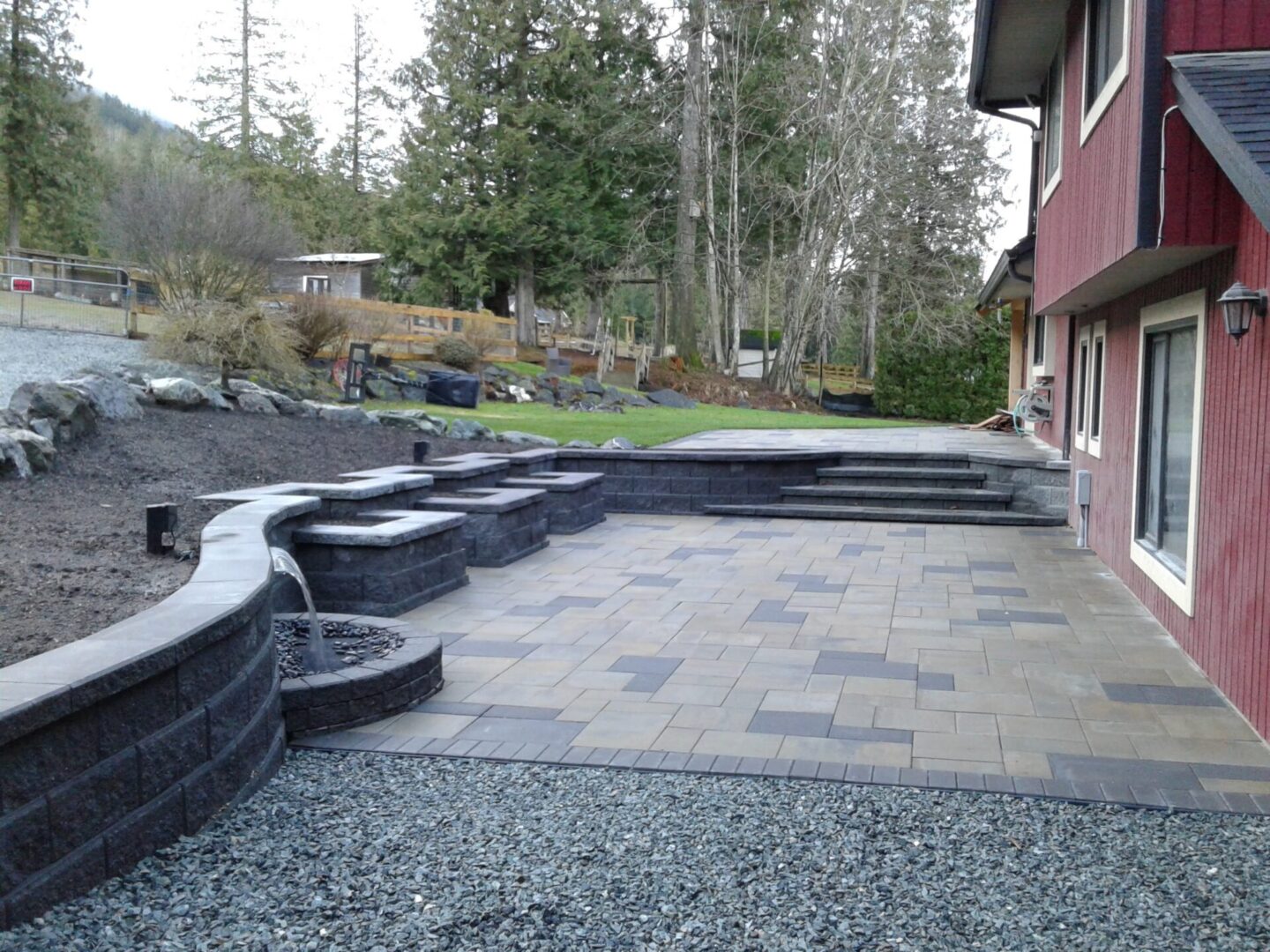 Paved patio with curved stone walls near a red house, overlooking a landscaped garden with trees.
