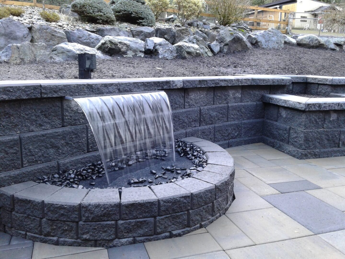 A modern outdoor water feature with water spilling from a metal chute into a curved stone basin surrounded by rocks and pebbles.