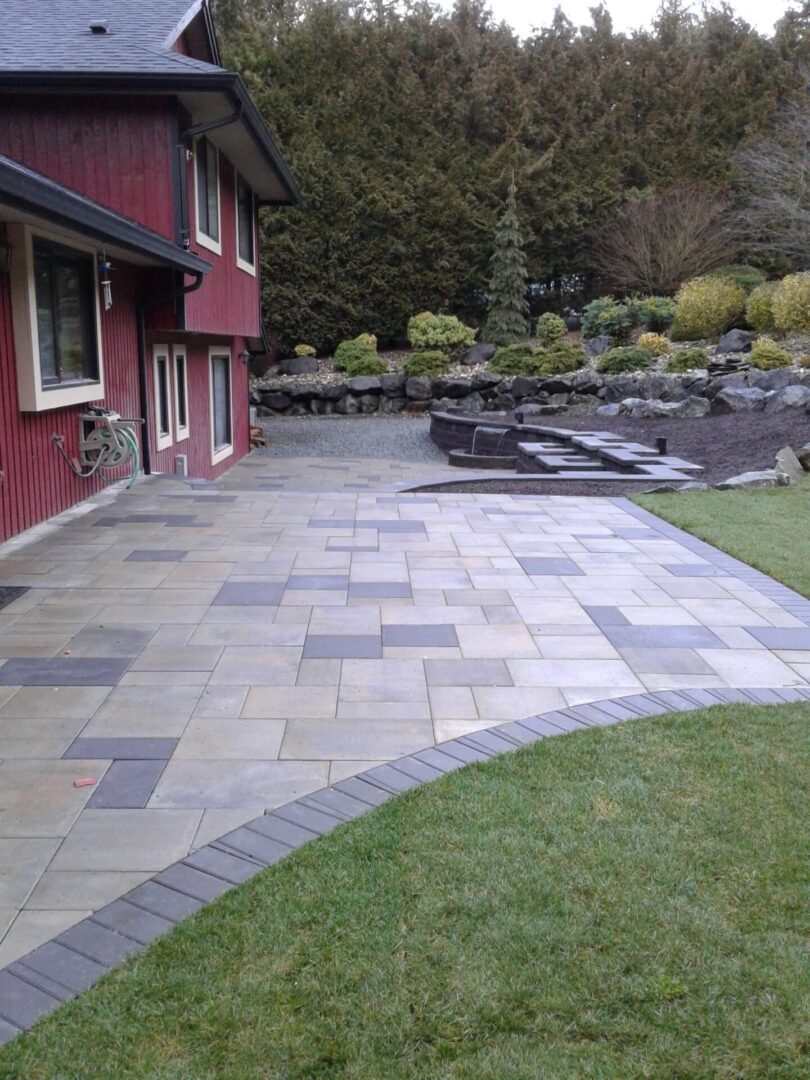 Newly installed slate tile patio next to a red house with landscaped garden and stone walls in the background.