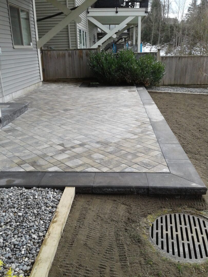Newly laid rectangular and square stone patio surrounded by fresh soil, gravel, and a wooden plank barrier, with a residential building in the background.