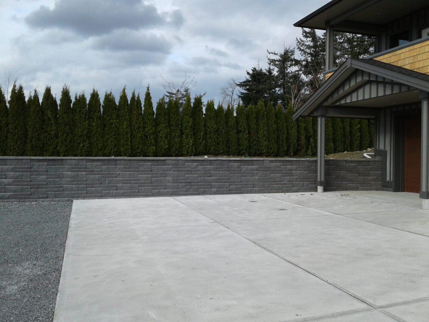 A wide driveway leading to a modern house with a gray stone wall and a row of tall, narrow evergreen trees under a cloudy sky.