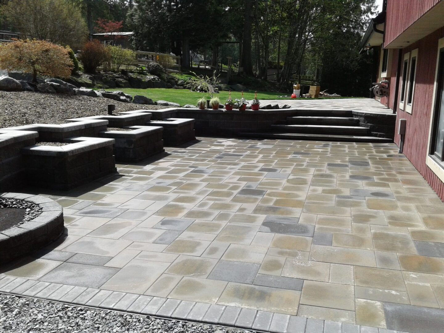 A neatly paved outdoor area with varying shades of rectangular stones, surrounded by green grass and multiple stone steps leading to a garden.