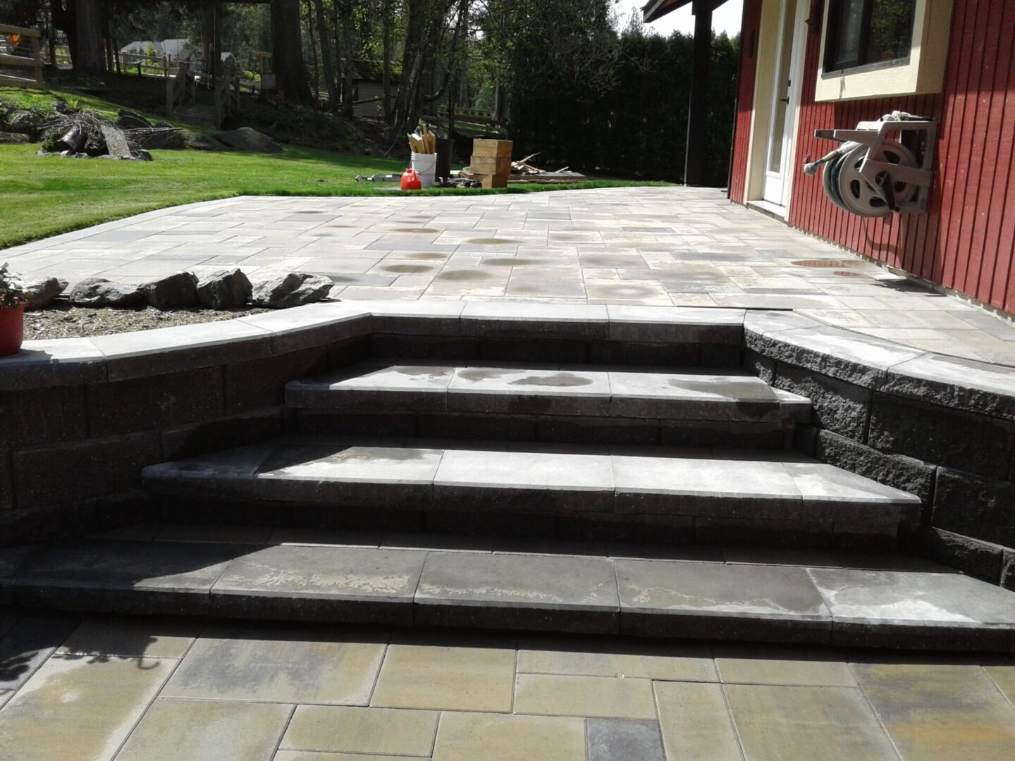 Paved backyard patio featuring grey stone slabs with a set of three steps, adjacent to a red building and a green lawn.