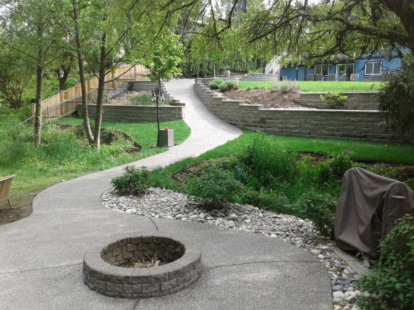 A winding concrete pathway through a landscaped garden with green trees, grass, and a small stone-lined pit in the foreground.