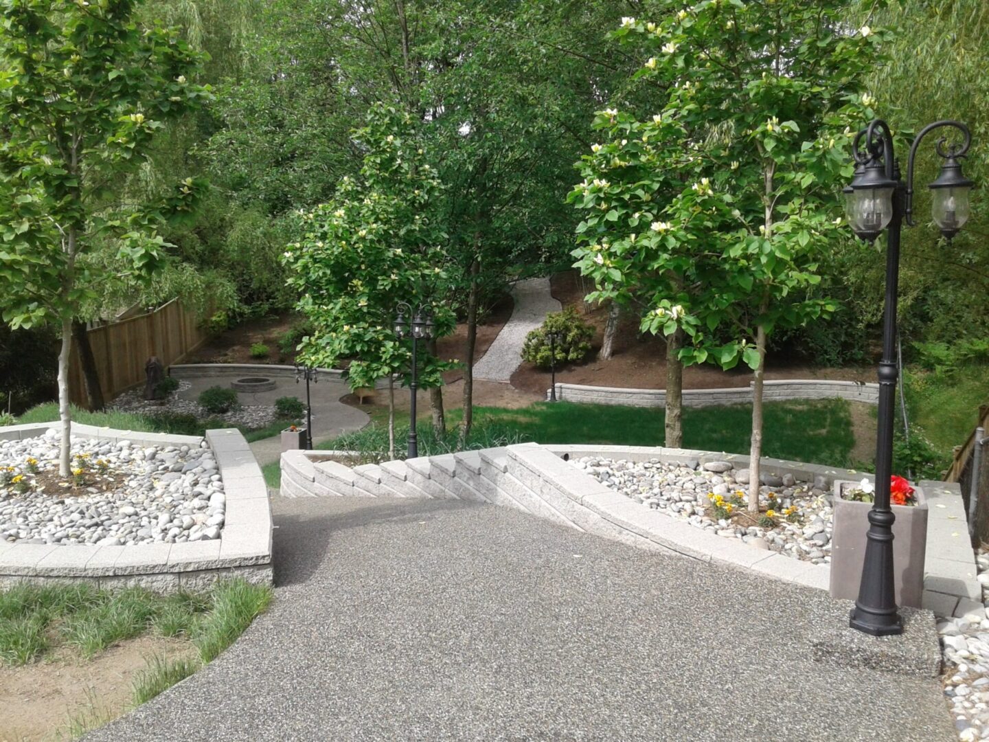 A paved pathway winding through a tranquil garden with young trees, white pebble borders, and lamp posts.