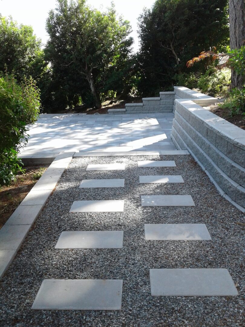 A garden pathway made of gravel with rectangular stepping stones, flanked by low stone walls and surrounded by lush greenery.