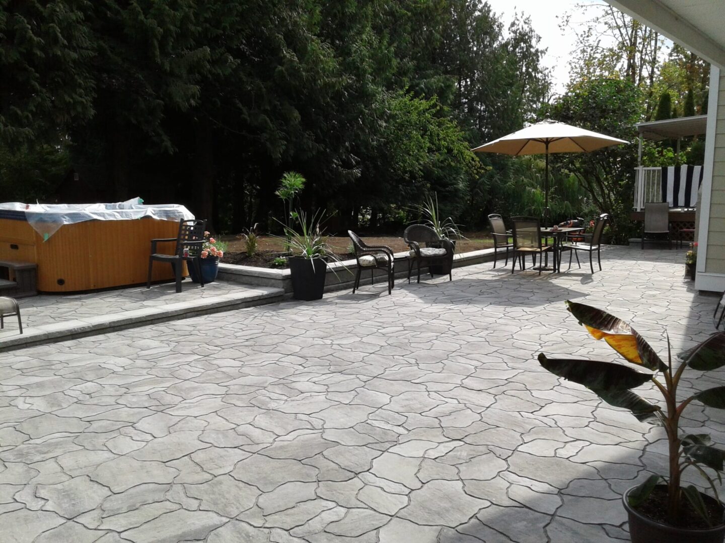 A spacious backyard patio featuring a hot tub, umbrella-shaded table, and scattered potted plants, set against dense greenery.