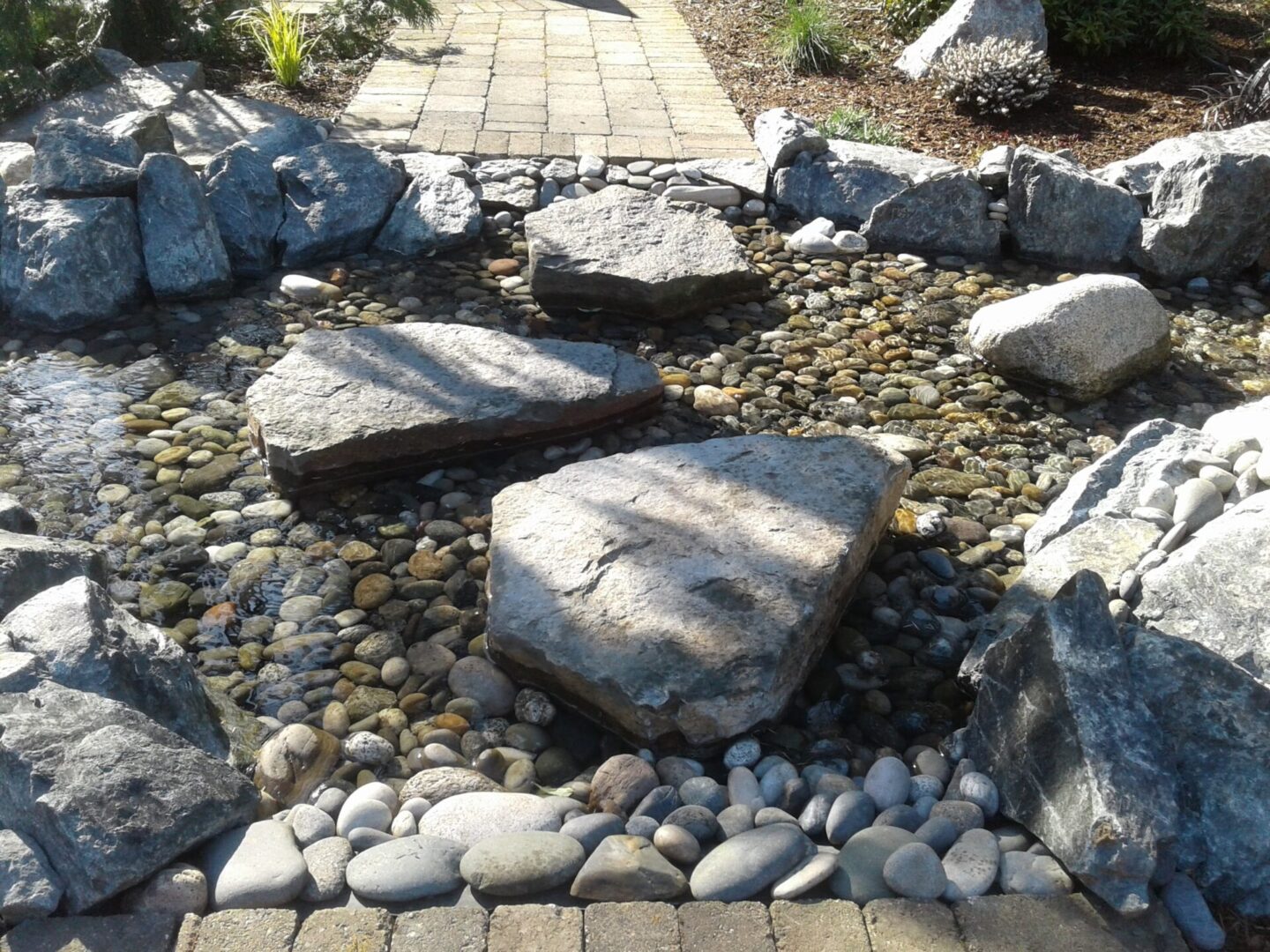 A landscape design featuring large stone slabs forming a stepping path across a bed of smooth, multicolored pebbles with surrounding greenery.