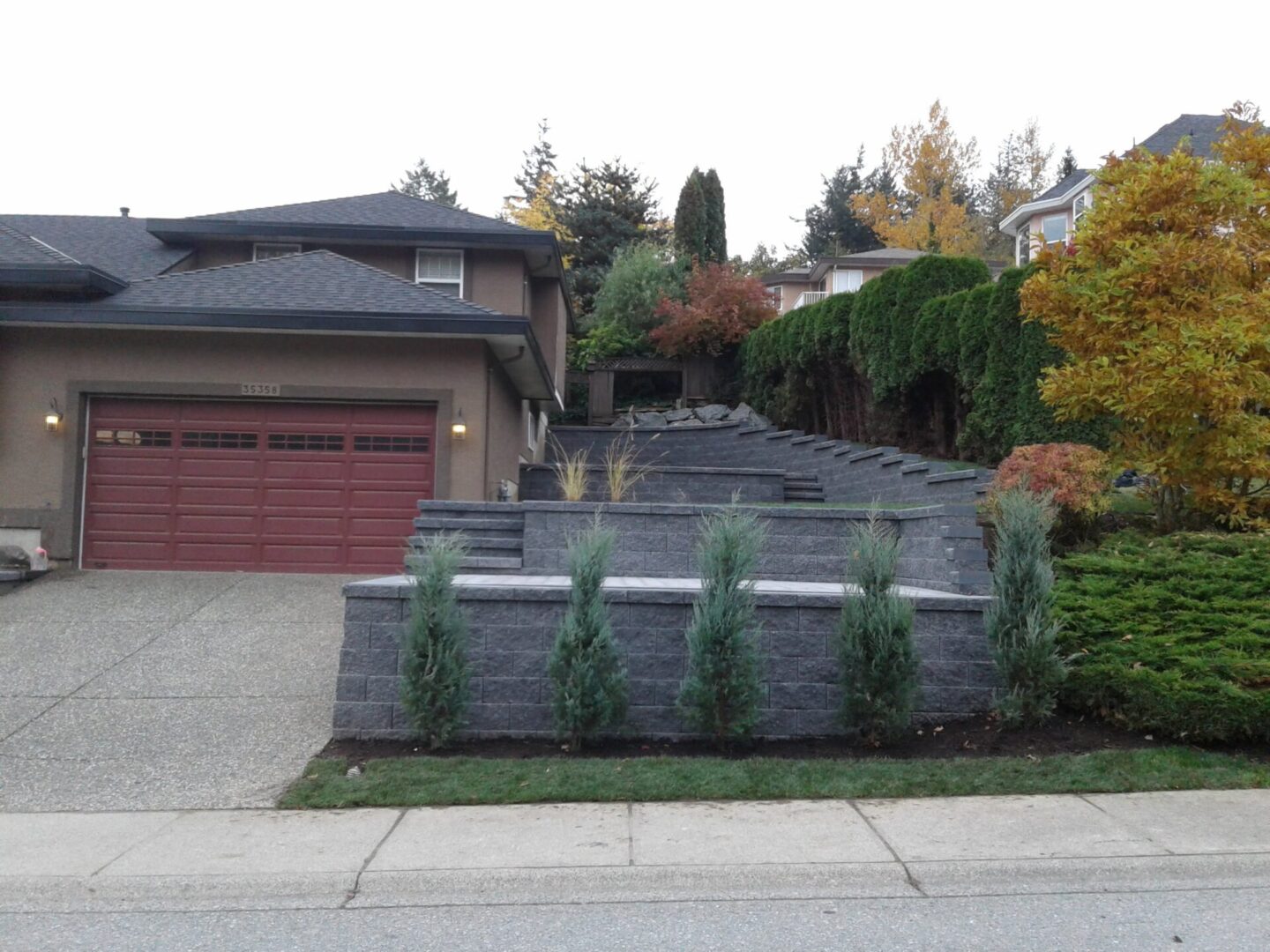 A residential house with a gray façade and double garage under a sloping roof, featuring a landscaped front yard with tiered stone walls and steps, surrounded by various shrubs and trees.