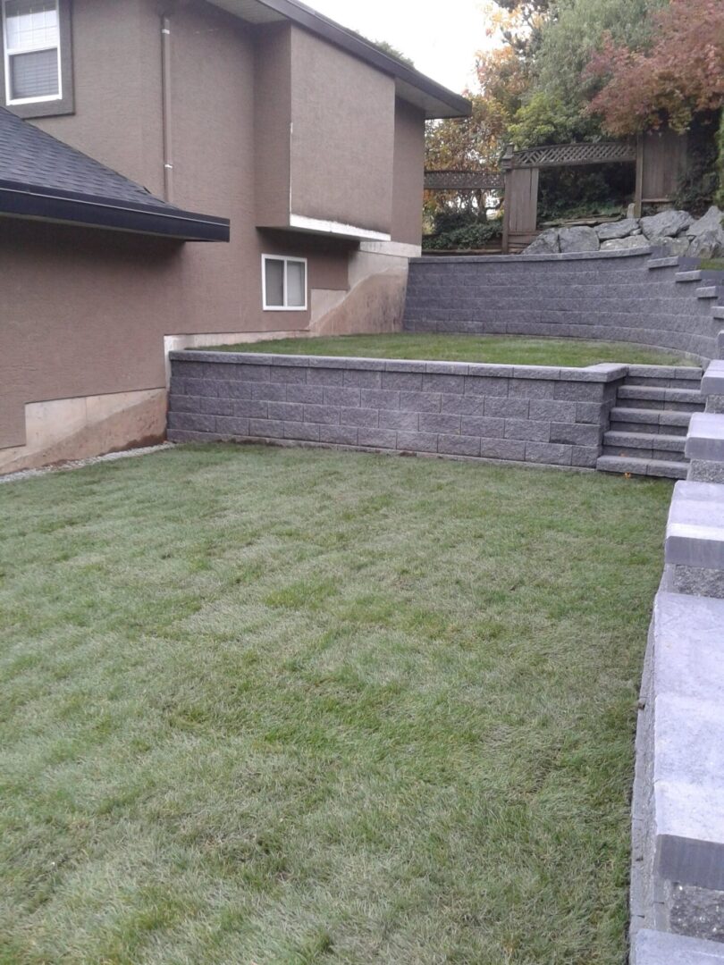 A well-manicured lawn bordered by a new gray stone retaining wall, with steps leading to a higher level beside a house.