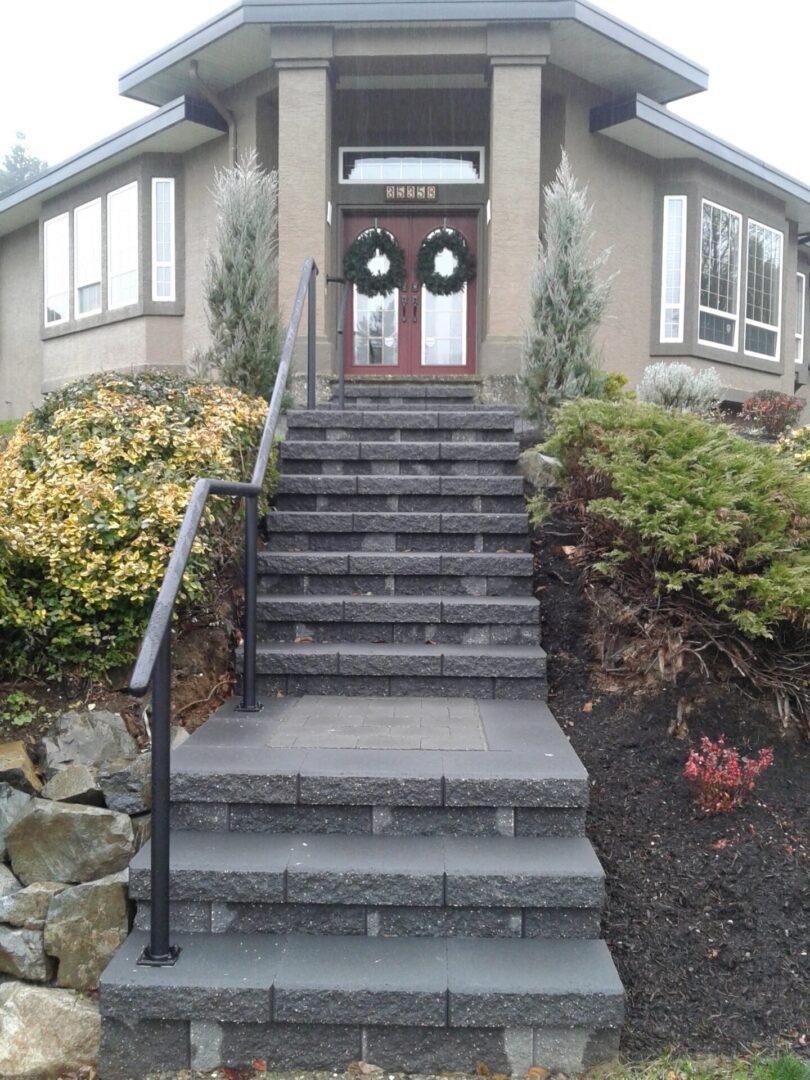 Front view of a house with a stone staircase leading to double doors with wreaths, flanked by tall grasses and shrubs.