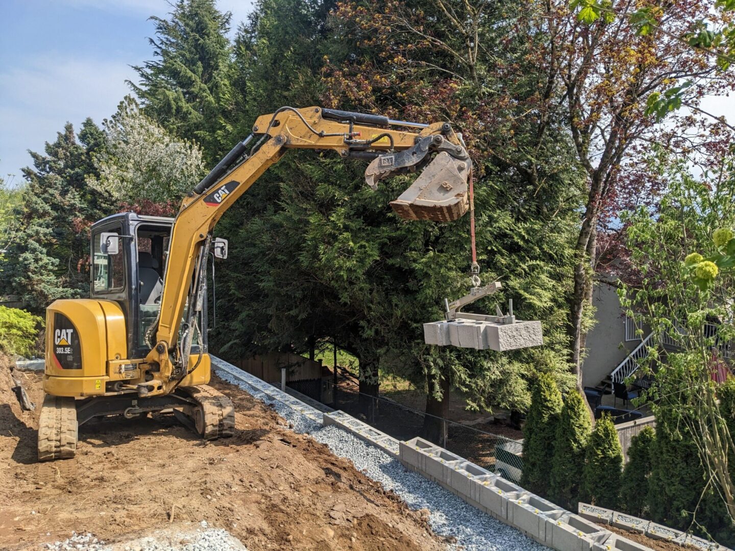 A yellow excavator moving landscaping bricks on a sloped yard with green trees and a house in the background.