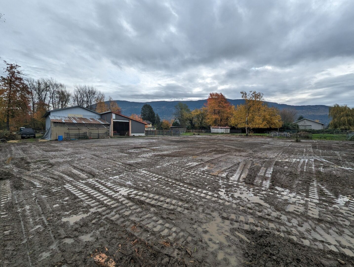 Muddy farmland with tire tracks, featuring barns and leafy trees under a cloudy sky with distant mountains.