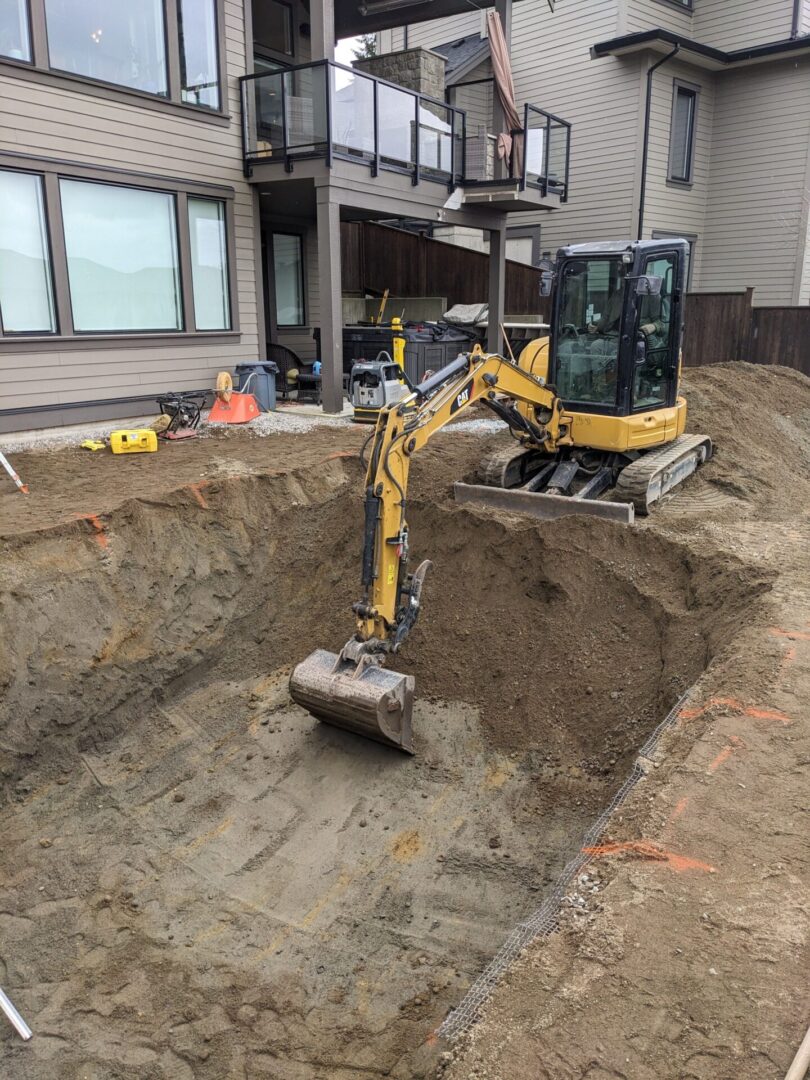 A yellow excavator digging in a large pit with marked boundaries next to a residential building under overcast skies.