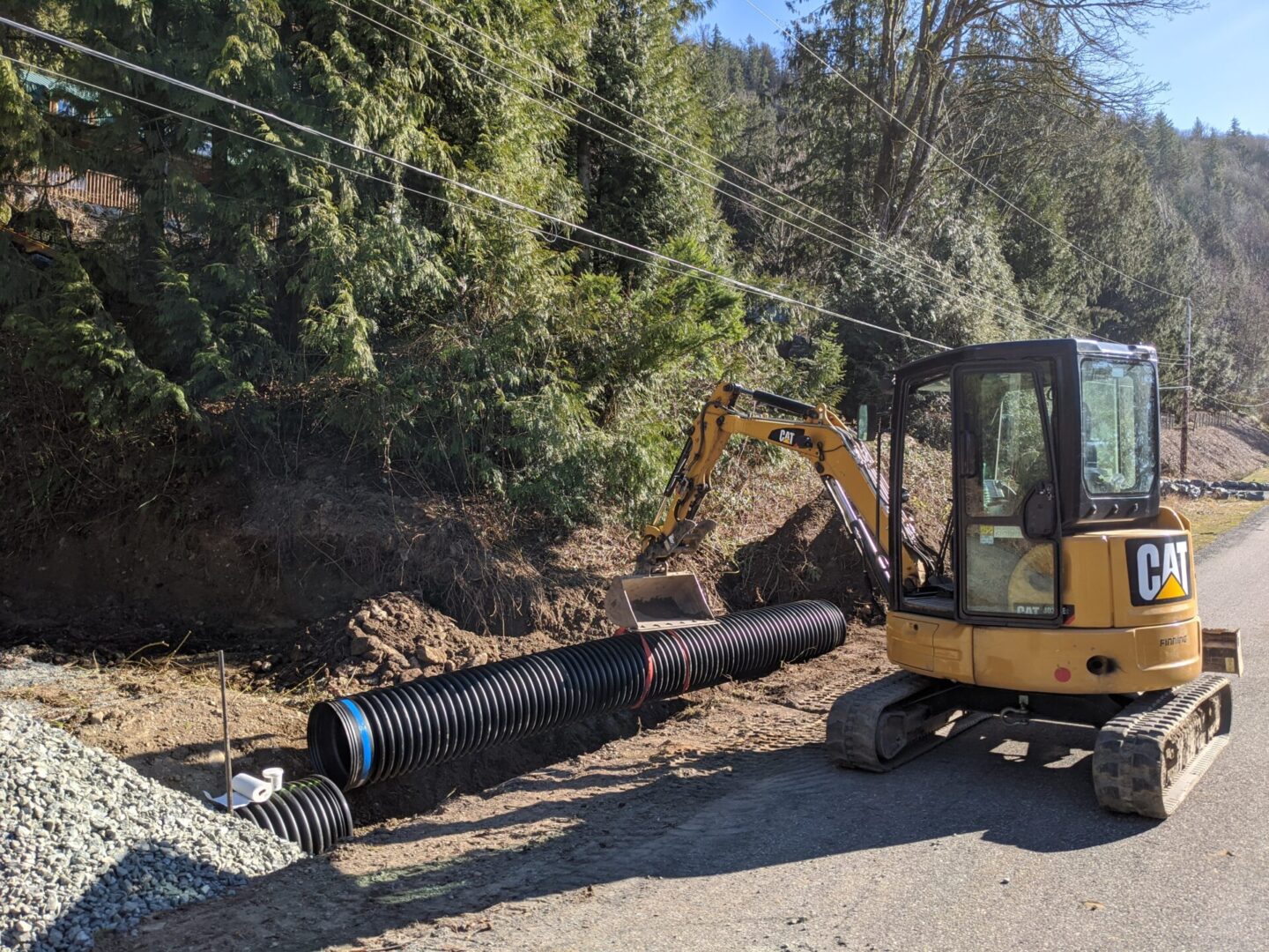 A yellow excavator installing large black drainage pipes beside a road, with a pile of gravel nearby and a forested hill in the background.