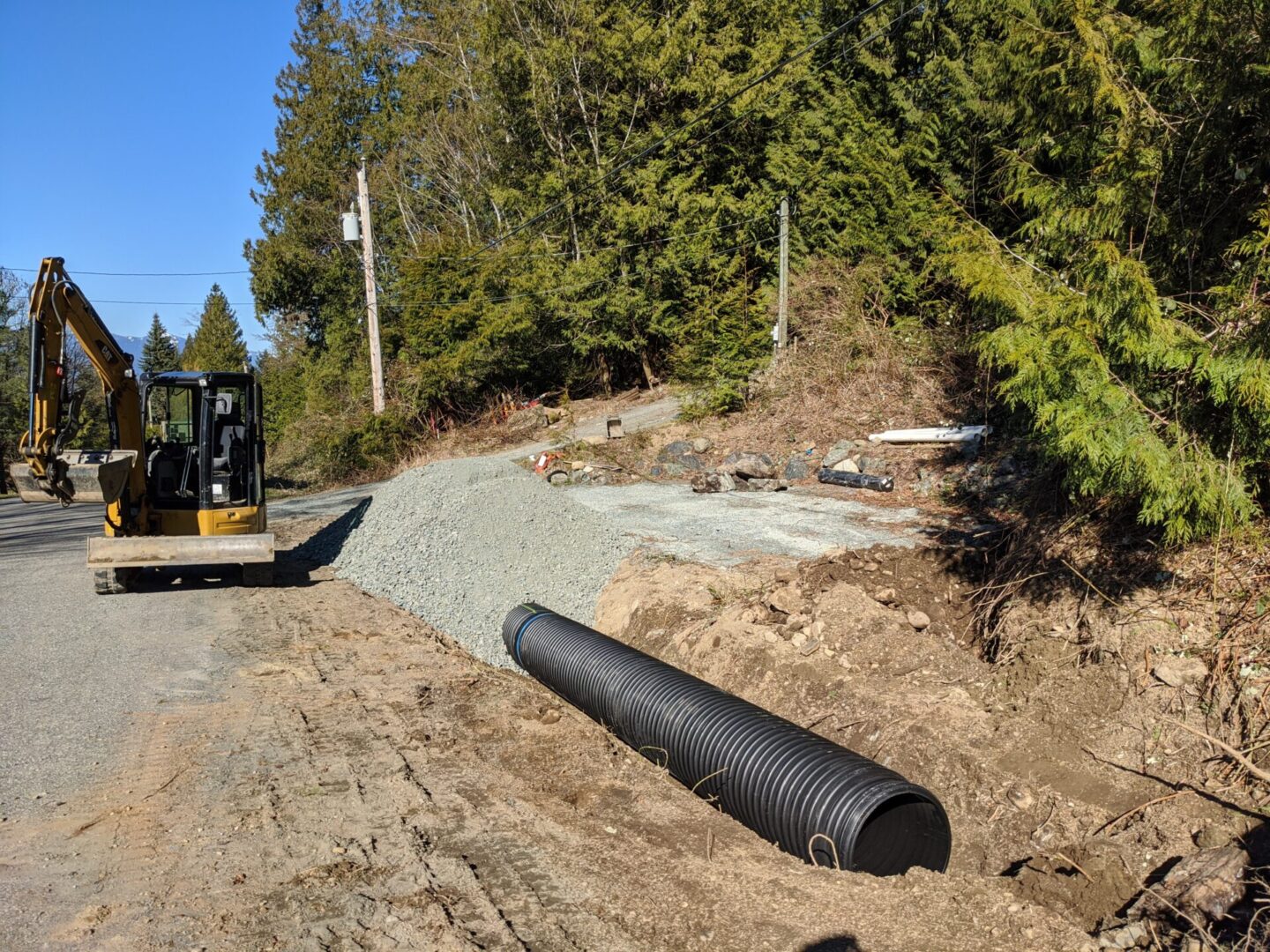 A construction site with a backhoe, a pile of gravel, and a large black drainage pipe next to a road in a wooded area.