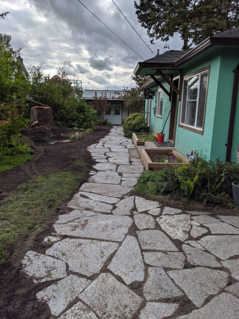 A pathway made of irregularly shaped stones leading to a green house with a small garden on the side under a cloudy sky.