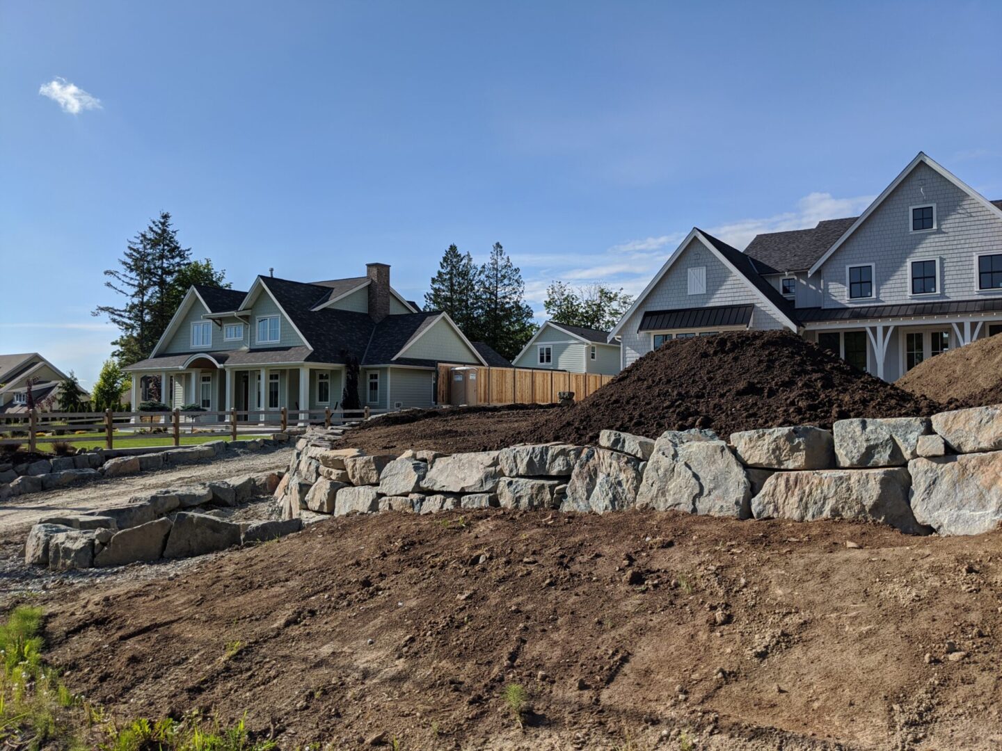 Suburban construction site with unfinished landscaping, featuring large piles of earth and boulders with houses in the background under a clear sky.