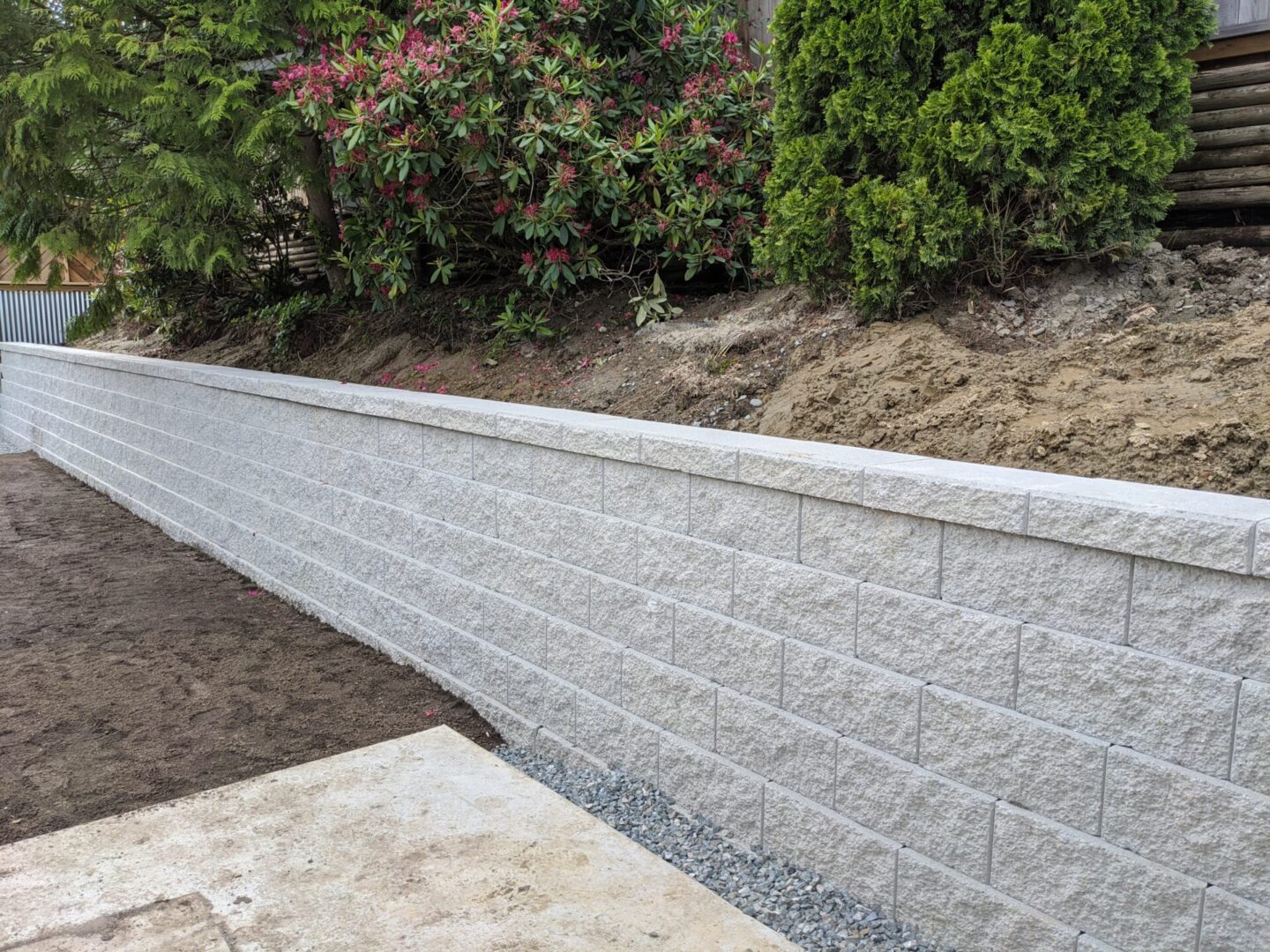 A newly constructed gray brick retaining wall separating a dirt area from a garden with shrubs and flowering plants.
