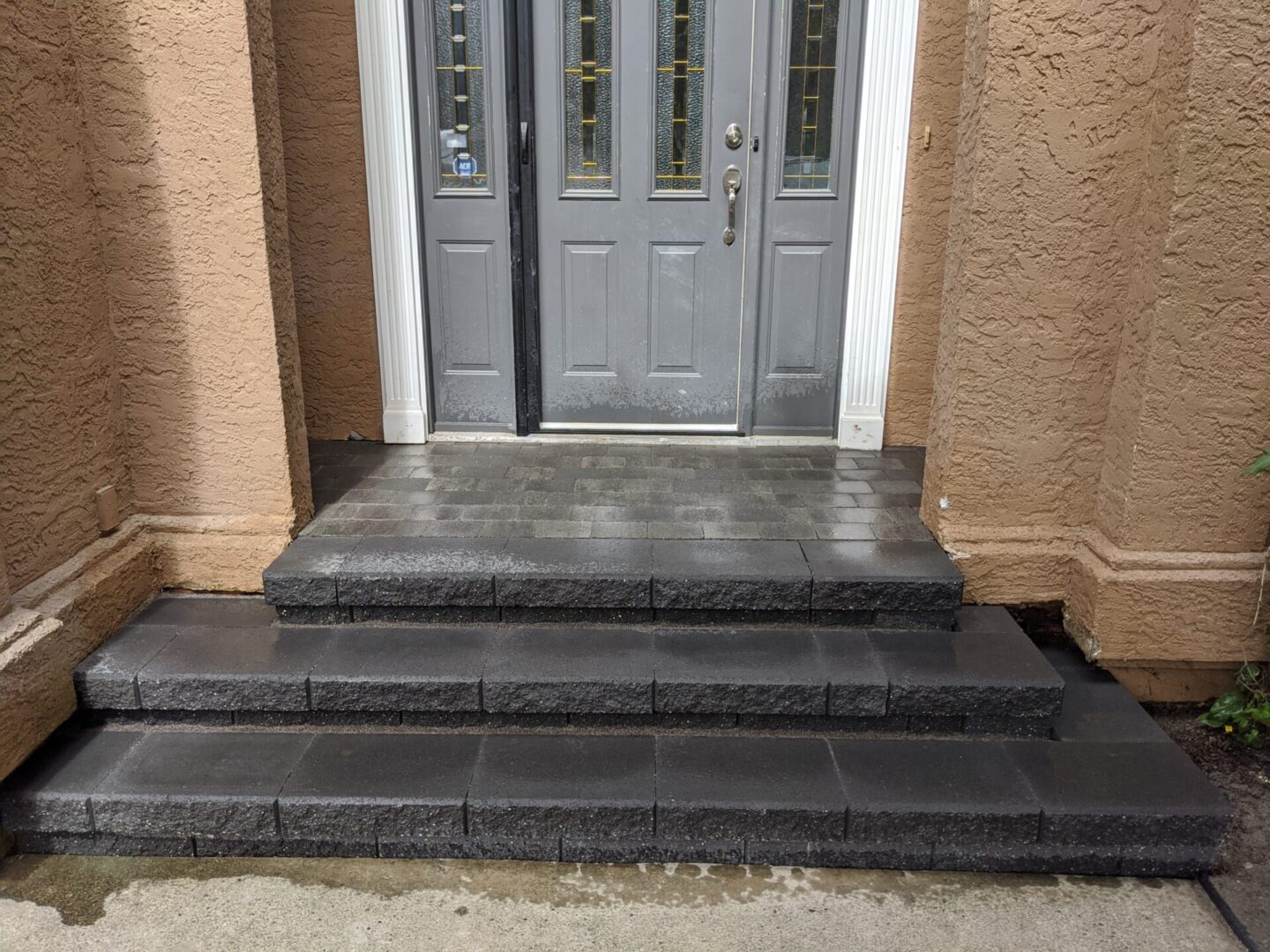 Gray double doors with glass panels set in a textured beige wall, above a wet black stone staircase with four steps.