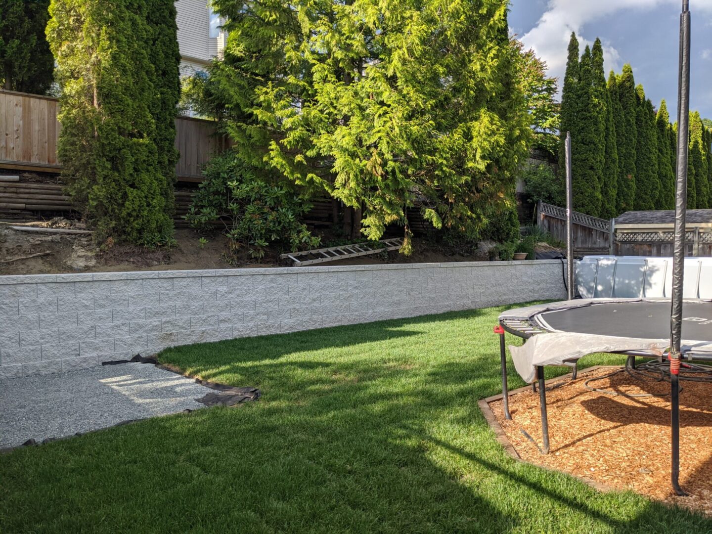 A well-maintained backyard featuring a grassy lawn, a trampoline on wood chips, surrounded by trees and a white fence under bright sunlight.