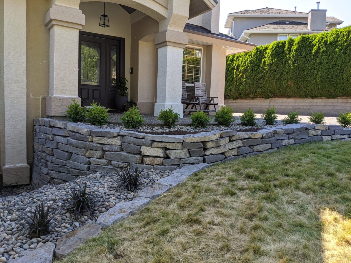 Front yard of a house with a stone retaining wall, landscaped garden, and a chair by the door.
