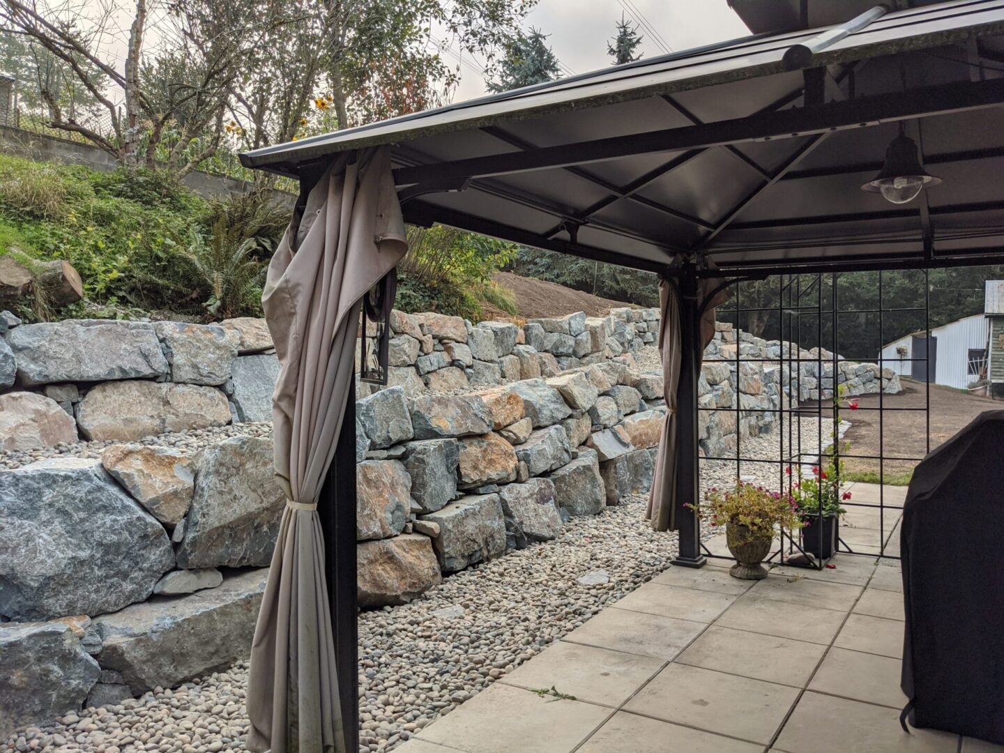 A gazebo with draped curtains on a gravel patio, adjacent to a rock retaining wall with plants and trees in the background.