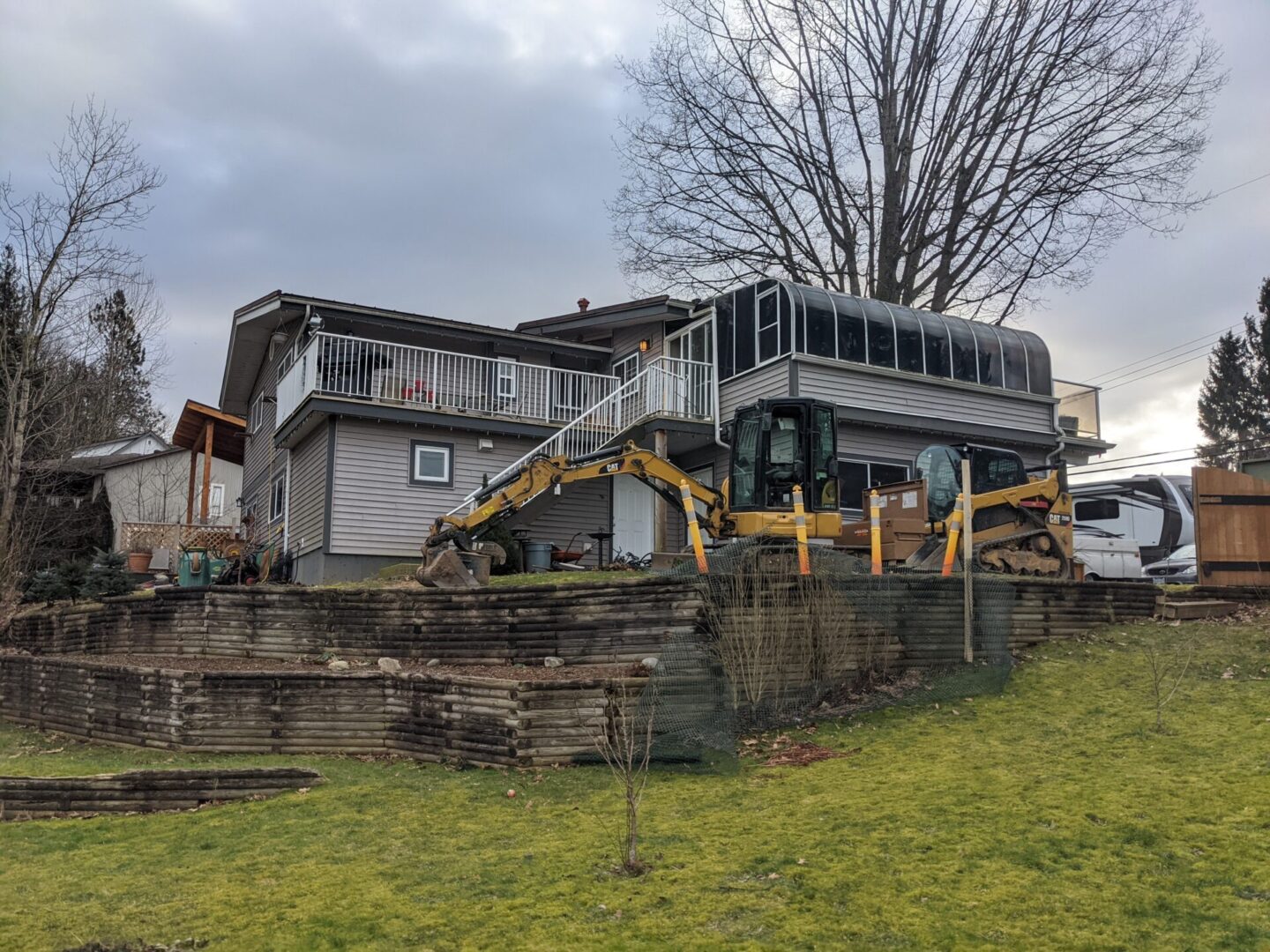 Backyard of a house with an excavator parked on grass, featuring tiered garden beds and a large tree. the house has a balcony and an enclosed porch.