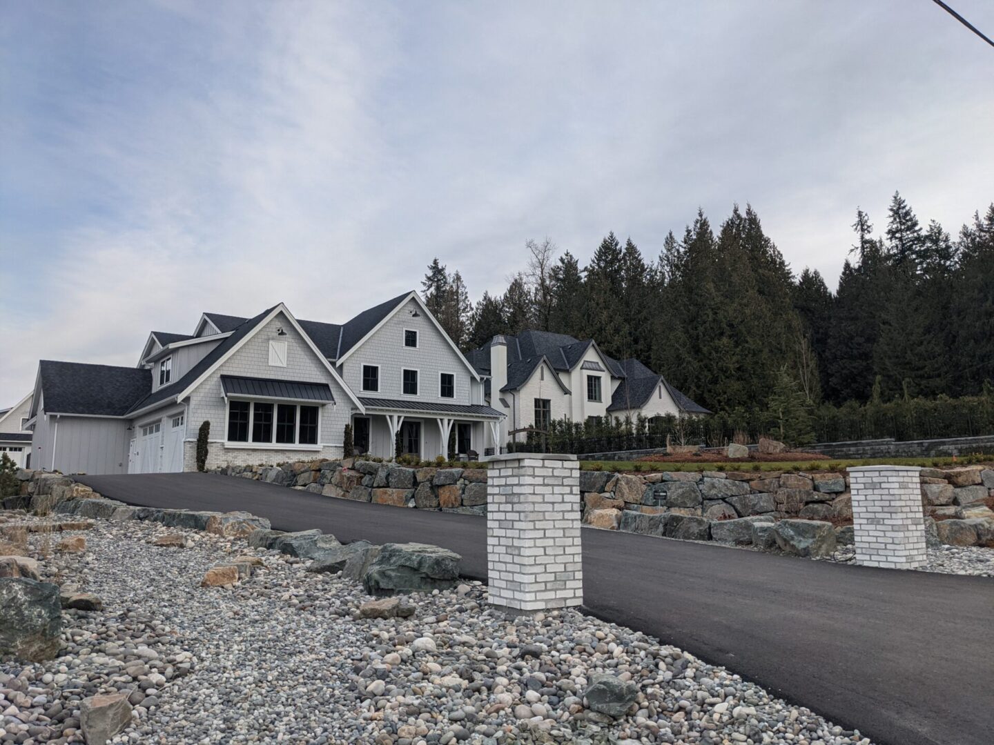 Large white house with multiple gables and a covered entrance, flanked by stone pillars and a pebble driveway, set against a backdrop of trees under a cloudy sky.