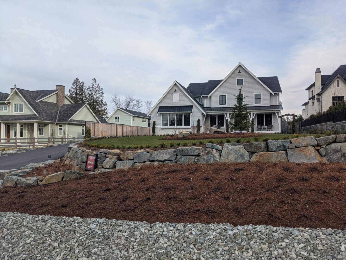 Newly landscaped suburban area with mulch and stone retaining wall, featuring a large white house and adjacent structures under an overcast sky.