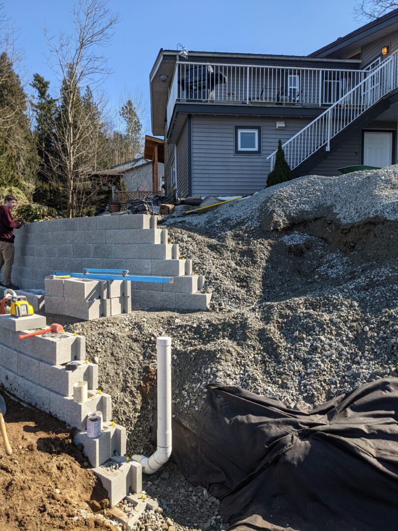 Residential construction site showing the early stages of a retaining wall being built, with piles of gravel, concrete blocks, and a house in the background.