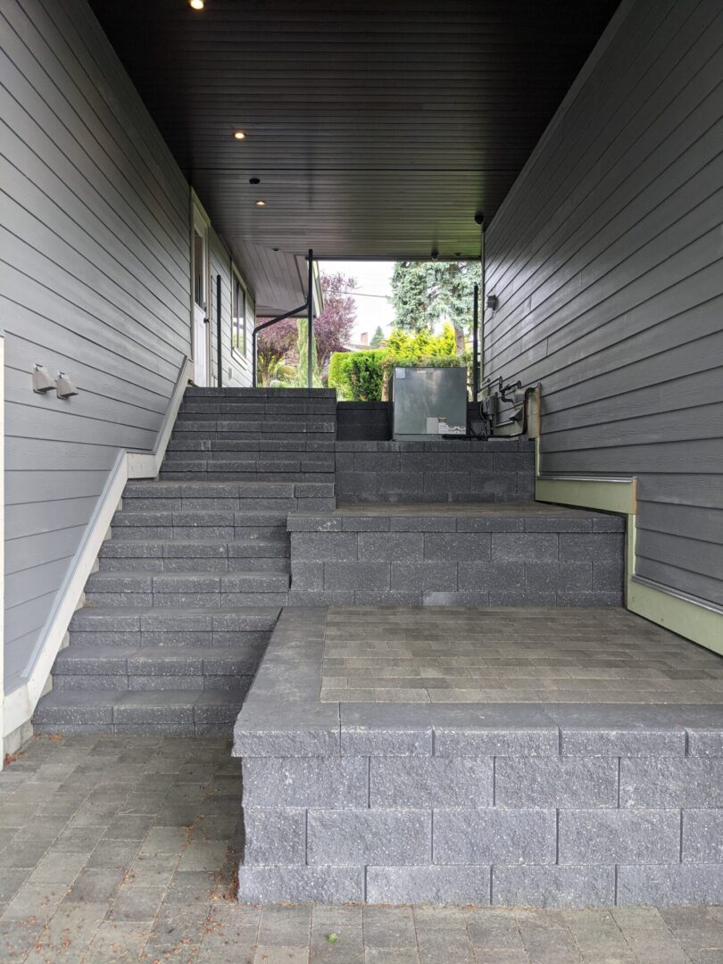 Gray stone staircase with multiple landings between gray-sided buildings, leading to a backyard with green foliage.