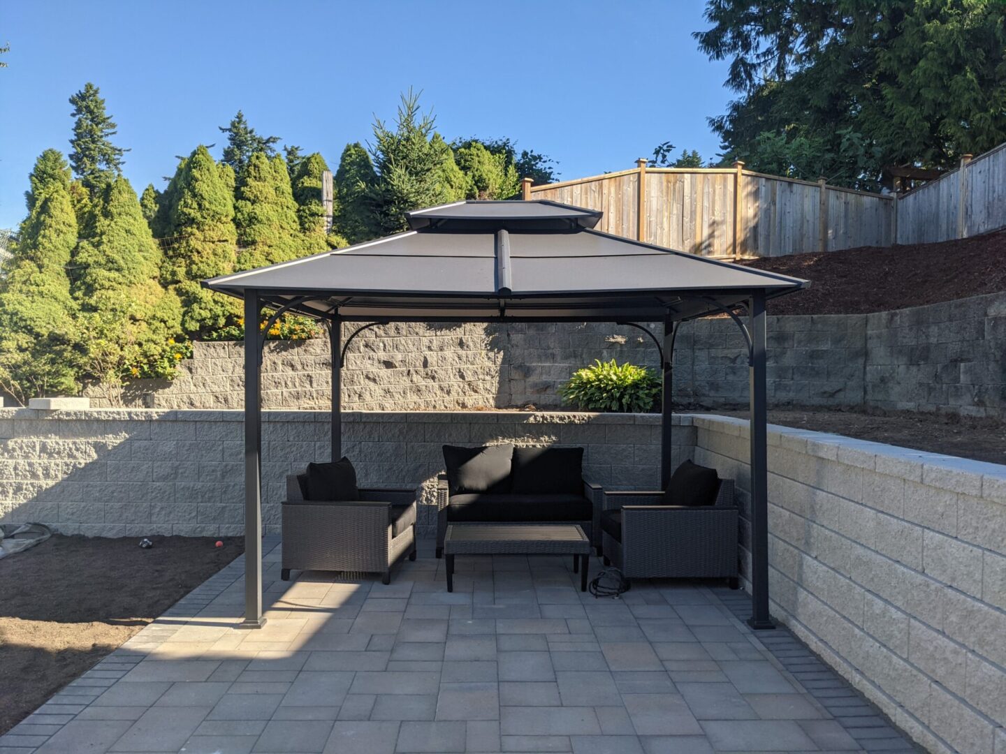 A modern backyard featuring a gazebo with a black sofa set on paved flooring, surrounded by retaining walls and trees under a clear sky.