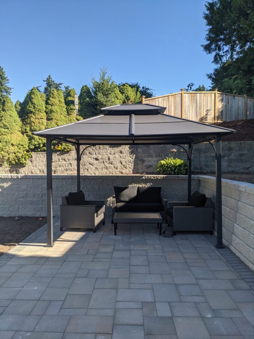 A patio with a black gazebo covering a seating area that includes a sofa and two chairs, set against a stone wall and trees under a clear blue sky.