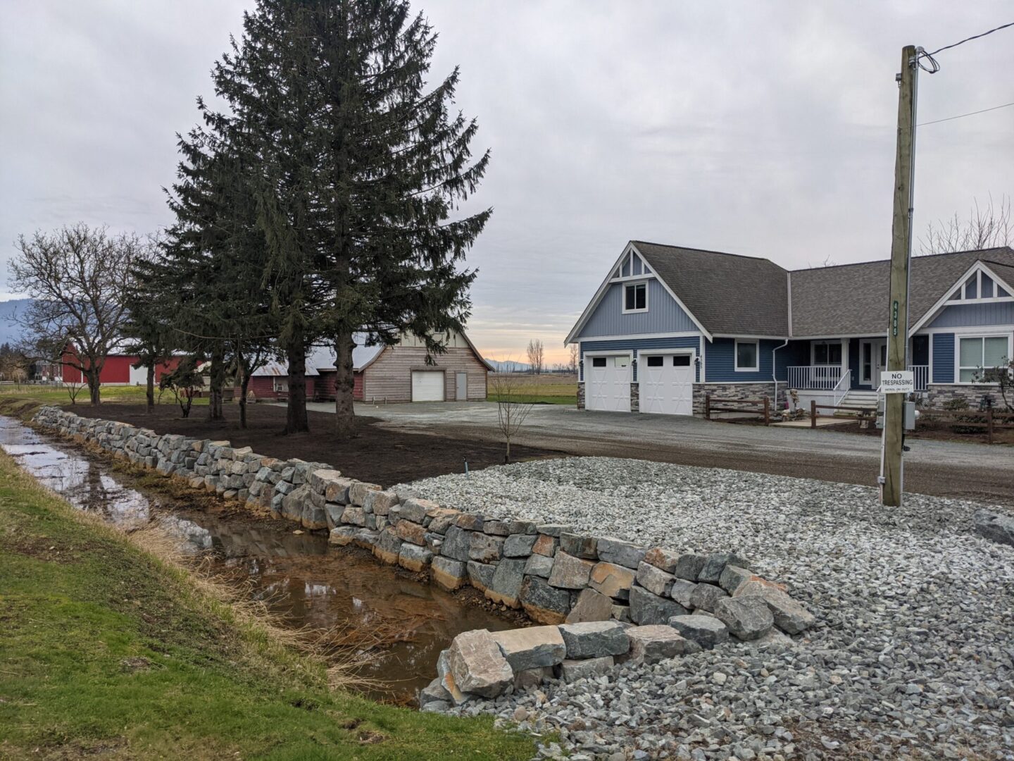 A rural landscape featuring a blue house with a white porch, a red barn in the distance, and a gravel-lined drainage ditch beside a road.