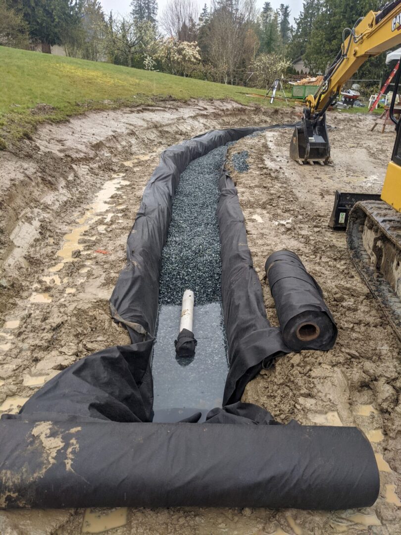 A construction site with a newly installed drainage system using geotextile fabric and gravel, surrounded by muddy ground and a backhoe in the background.