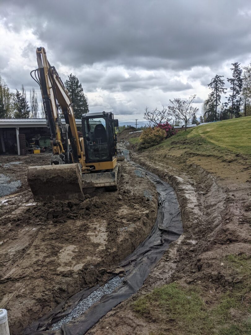 An excavator digging a curving trench in a muddy yard, lined with a black fabric, under a cloudy sky.