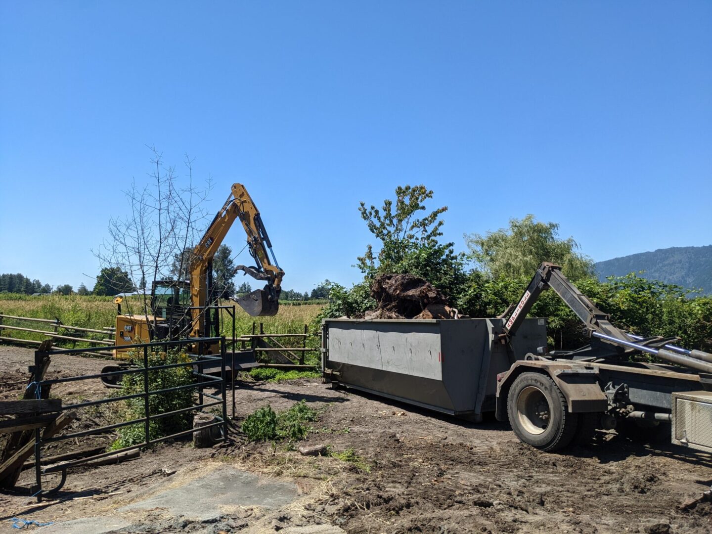 An excavator loading soil into a dump truck in a rural setting with trees and blue sky in the background.