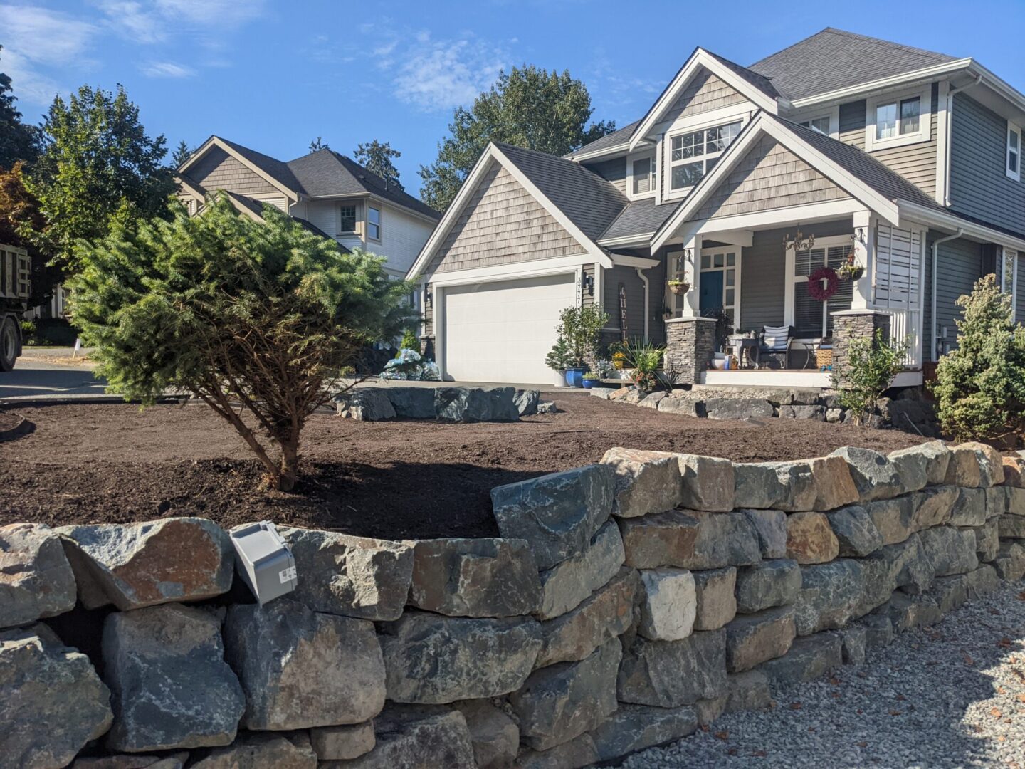 A suburban street with well-manicured homes, featuring a stone wall and fresh landscaping in the foreground.