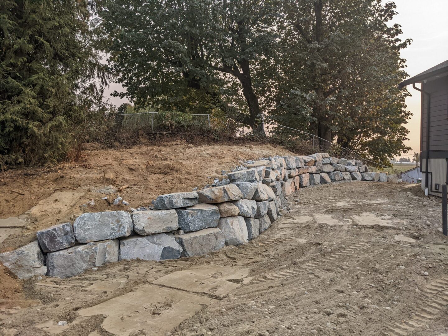 A newly constructed grey stone retaining wall on a dirt embankment next to a building, with trees in the background under a hazy sky.