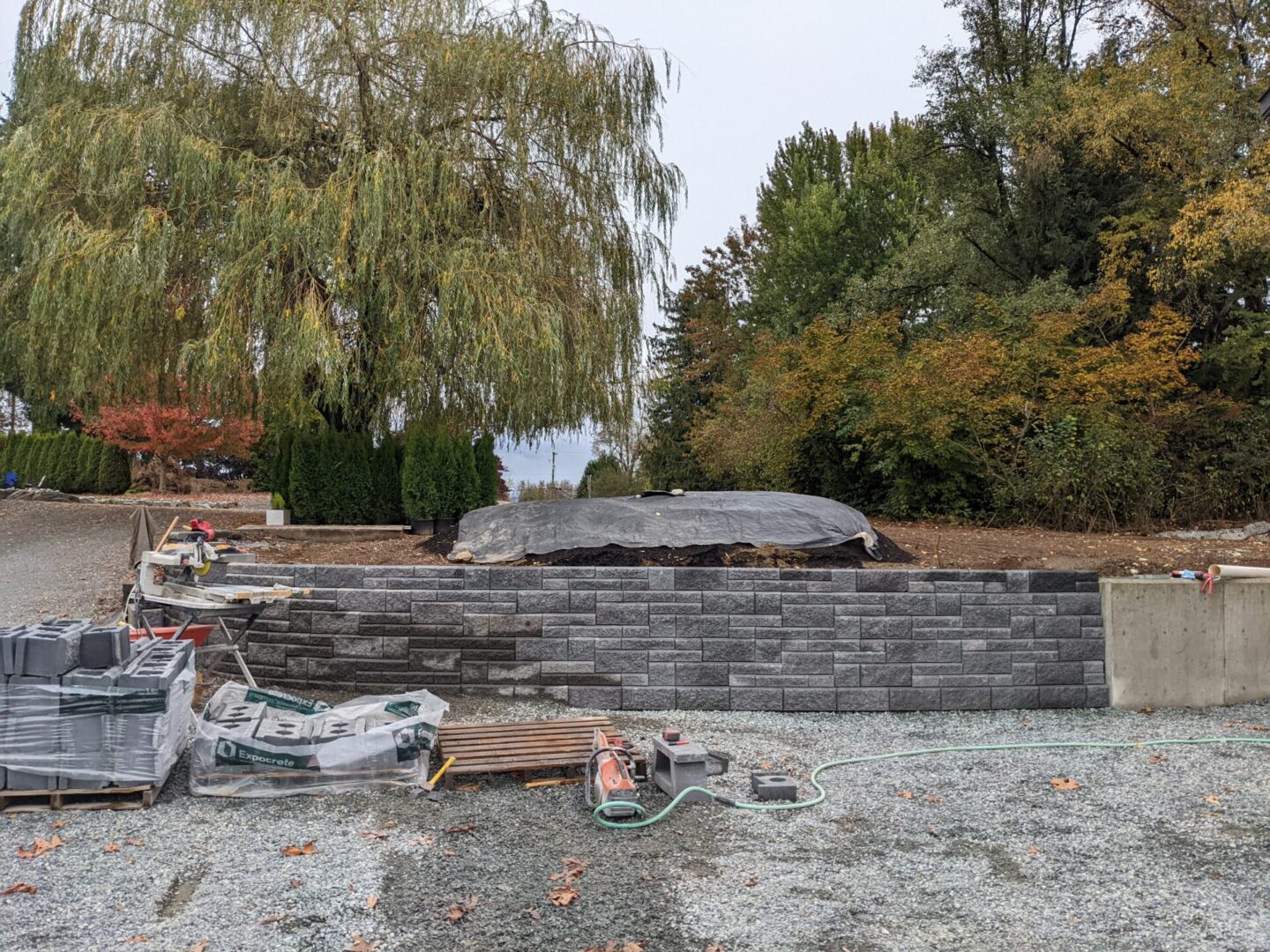 Construction site with stone retaining walls under development, building materials scattered around, and a weeping willow in the background.