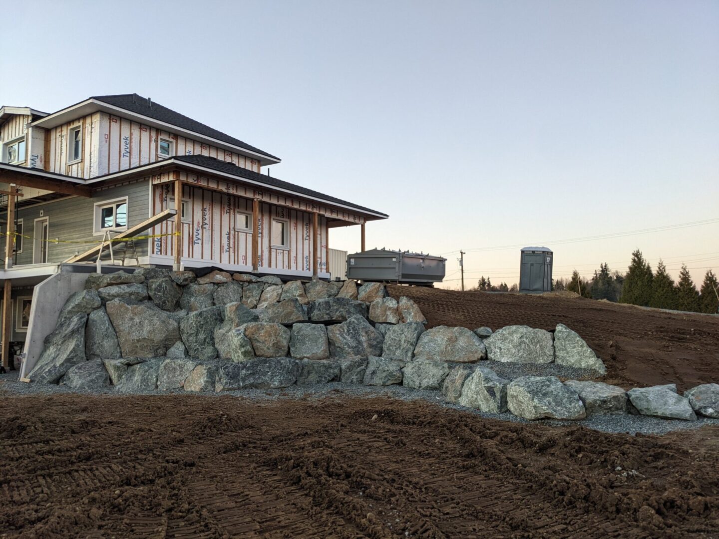 A new house with exposed insulation under construction, featuring a stone retaining wall in the foreground and a portable toilet on the side.