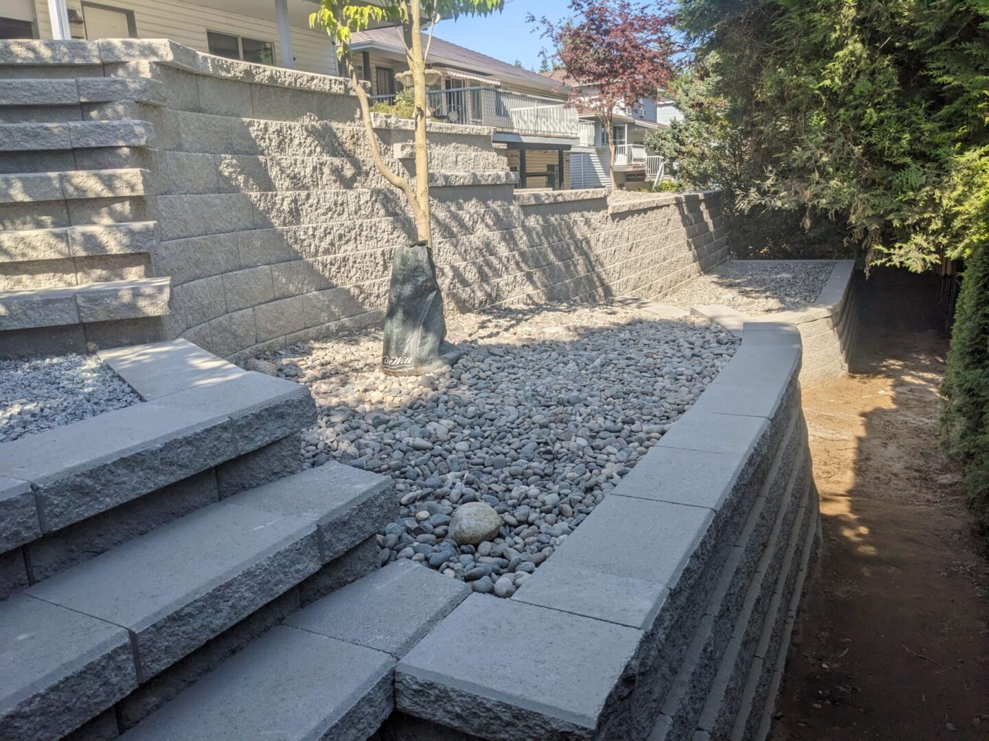 A residential outdoor area with structured gray stone steps and walls, landscaped with gravel and small trees, in a sunny setting.