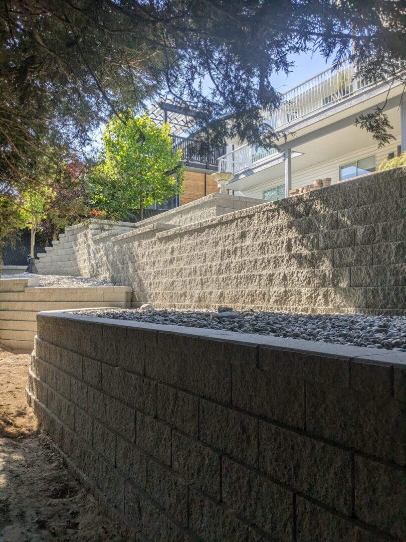 Stone retaining wall with stairs leading to a house with a balcony, surrounded by trees and gravel on the ground, under clear daylight.