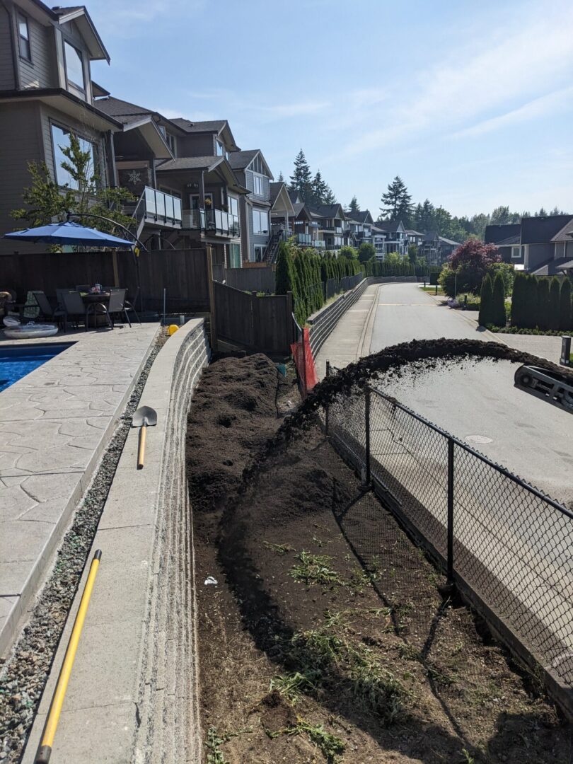 A curved chain-link fence damaged and partially collapsed into a trench alongside a residential street with houses and a clear sky.