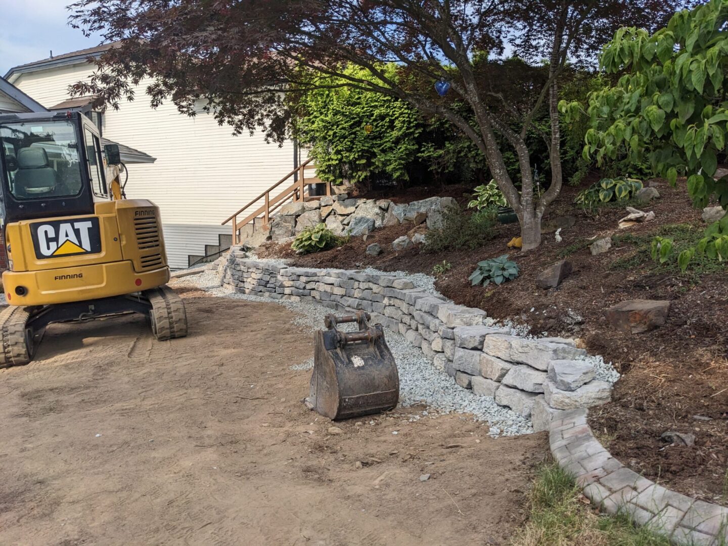 A construction site with a yellow cat excavator near a newly built stone retaining wall and landscaping in a residential area.