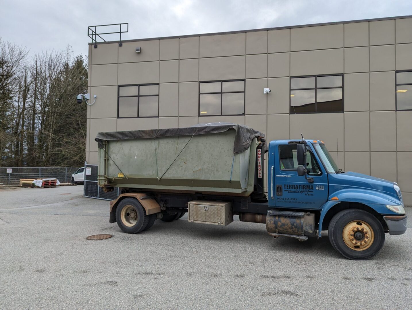 A blue dump truck with a dirty green bin parked beside a gray industrial building under a cloudy sky.