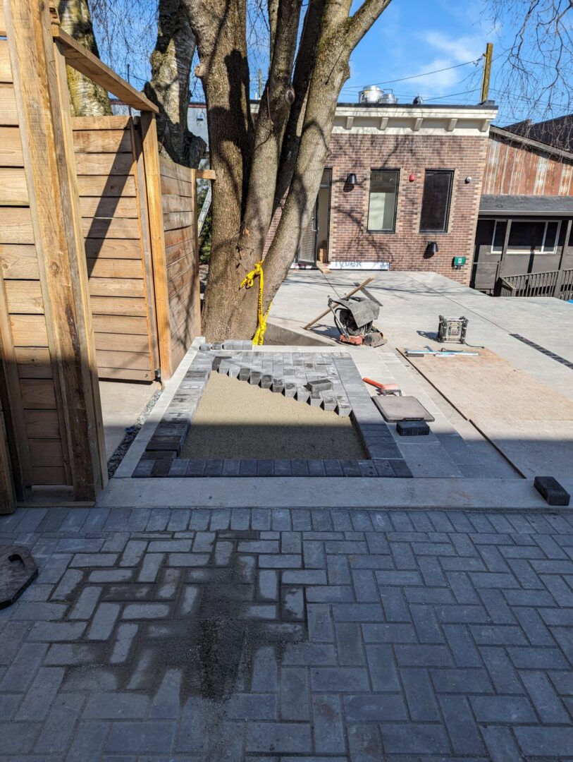 Unfinished backyard patio construction with new pavers and sand base, tools scattered around, next to a large tree and wooden structure.